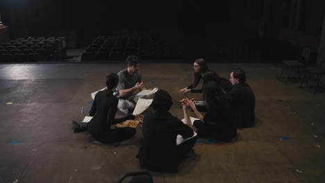 Happy-group-of-actors-holding-script-sheets-in-their-hands-and-actively-discussing-their-roles-with-the-director-on-stage-in-the-theater-during-rehearsal