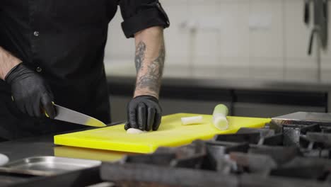 Primer-Plano-De-Un-Chef-Profesional-Con-Uniforme-Negro-Y-Guantes-De-Goma-Corta-Puerros-En-Una-Tabla-De-Cortar-Amarilla-En-La-Cocina-De-Un-Restaurante.-Un-Chef-Experimentado-Corta-Verduras-Con-Destreza-Mientras-Prepara-Un-Plato-En-La-Cocina.