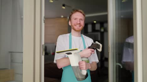 Portrait-of-a-confident-blond-man-with-a-beard-in-a-white-T-shirt-and-with-a-blue-apron-a-cleaner-holds-in-his-hands-two-vacuum-cleaners-for-windows-during-his-cleaning-in-a-modern-apartment-on-call-from-a-cleaning-company