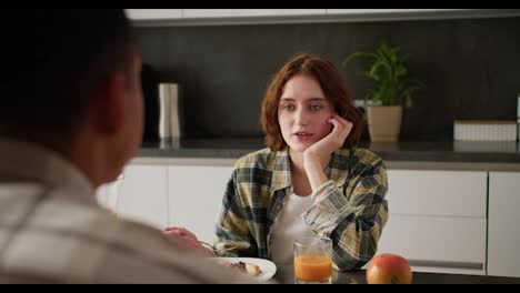 Over-the-shoulder-a-young-adult-girl-with-brown-hair-and-a-bob-hairstyle-communicates-with-her-boyfriend-with-Black-skin-color-during-their-breakfast-together-in-a-modern-kitchen-in-an-apartment