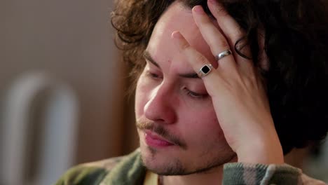 Close-up-of-a-sad-brunette-guy-with-curly-hair-holds-his-head-with-his-hand-and-thinks-hard-at-home-in-a-modern-apartment