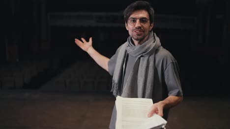 Portrait-of-a-confident-young-brunette-man-in-glasses-of-a-confident-stage-director-in-a-gray-T-shirt-and-a-gray-scarf-with-a-script-in-his-hands-on-stage-in-the-theater