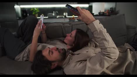 A-brunette-man-with-stubble-and-his-brunette-girlfriend-in-a-beige-T-shirt-are-looking-at-something-in-their-smartphones-while-lying-on-a-gray-sofa-in-a-modern-studio-apartment