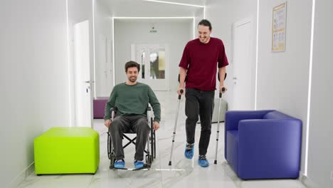 A-happy-brunette-man-with-stubble-in-a-green-jacket-rides-on-a-chair-for-the-disabled-during-his-rehabilitation-and-communicates-with-a-man-in-a-red-T-shirt-who-is-walking-on-crutches-in-a-modern-clinic