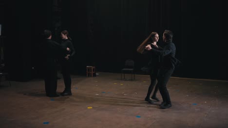 Two-pairs-of-confident-actors-in-black-suits-dance-on-stage-during-their-rehearsal-for-a-play-in-the-theater.-Careful-preparation-of-actors-in-black-suits-before-performing-and-dancing-in-the-theater