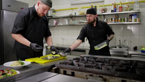 A-male-trainee-chef-in-a-black-uniform-prepares-a-dish-with-the-help-of-an-experienced-chef-in-the-kitchen-in-a-restaurant.-A-male-experienced-chef-helps-a-trainee-to-prepare-a-dish-correctly-and-use-the-right-ingredients-in-the-kitchen-in-a-restaurant