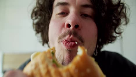 Close-up-portrait-of-a-Happy-guy-with-curly-hair-and-mustache-biting-a-burger-and-enjoying-it-in-the-morning-in-a-modern-apartment