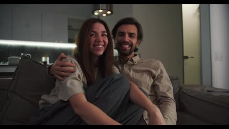 Portrait-of-a-happy-brunette-girl-in-a-beige-T-shirt-together-with-her-brunette-boyfriend-with-stubble-they-sit-on-the-sofa-and-laugh-in-a-modern-apartment-in-the-evening