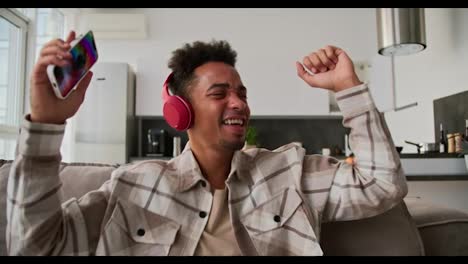 Happy-young-man-with-Black-skin-color-brunette-in-red-headphones-and-a-cream-plaid-shirt-listens-to-cheerful-music-along-with-his-phone-in-his-hands-while-sitting-on-a-brown-sofa-and-dancing-in-a-modern-apartment