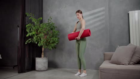 A-brunette-girl-in-a-gray-top-and-green-sweatpants-lays-out-a-red-mat-and-begins-her-exercise-in-a-modern-apartment-at-home.-A-young-brunette-girl-in-a-sports-summer-uniform-does-sports-on-the-red-carpet-near-a-houseplant-and-a-sofa-at-home