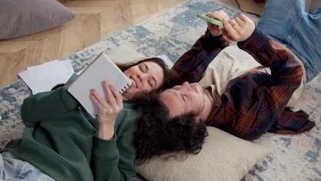 A-happy-brunette-girl-in-a-green-jacket-lies-on-the-floor-on-pillows-with-her-brunette-boyfriend-writes-something-in-a-notepad-while-the-guy-communicates-on-social-networks-using-a-smartphone-at-home