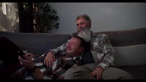 A-happy-brunette-man-in-a-checkered-shirt-lies-on-his-elderly-boyfriend-with-gray-hair-and-a-lush-and-white-beard-in-a-checkered-shirt-and-they-watch-TV-while-sitting-on-a-gray-sofa-in-a-modern-apartment