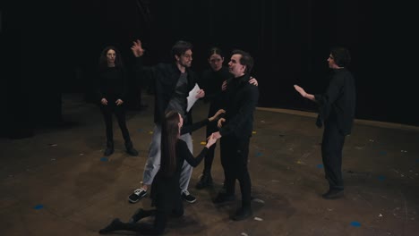 A-group-of-several-young-actors-in-black-suits-rehearse-their-roles-while-their-stage-director-communicates-with-them-and-portrays-their-roles-on-stage-in-the-theater