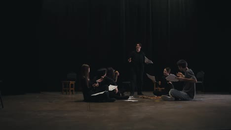 A-confident-young-brunette-guy-in-a-black-suit-holds-sheets-of-script-in-his-hands-and-plays-his-role-during-a-rehearsal-for-the-play-along-with-his-fellow-actors-and-the-director-on-stage