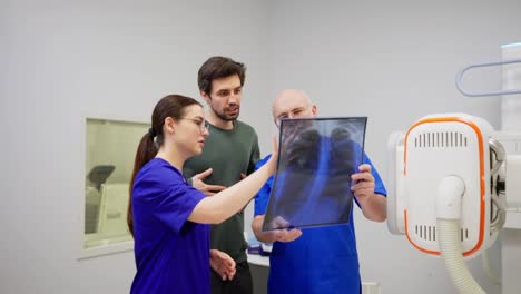 A-confident-doctor-in-glasses-with-a-beard-together-with-his-assistant-a-brunette-girl-in-a-blue-uniform-tells-the-brunette-guy-and-holds-a-chest-X-ray-in-front-of-them-in-a-modern-clinic-in-the-office