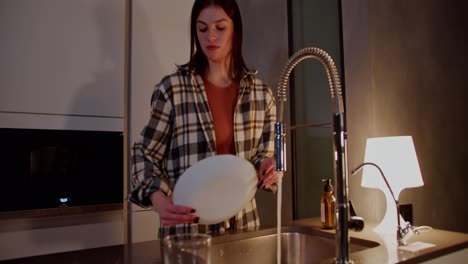 Confident-brunette-girl-in-a-checkered-shirt-washes-dishes-and-rinse-aid-with-a-sponge-and-cleaning-product-in-the-kitchen-in-the-evening-in-a-modern-apartment-routine