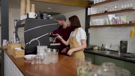 A-confident-male-barista-in-a-red-jacket-tells-a-brunette-girl-trainee-how-to-make-coffee-correctly-and-operate-a-coffee-machine-in-a-cafe