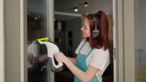 A-brunette-girl-in-black-wireless-headphones-and-a-white-T-shirt-in-a-blue-apron-as-a-cleaning-lady-washes-glass-doors-using-a-special-device-in-a-modern-apartment