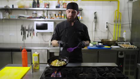 Confident-male-chef-in-a-black-uniform-with-a-black-bandana-fries-vegetables-on-a-gas-burner-turning-them-over-for-even-cooking-using-a-spatula-in-the-kitchen-in-a-restaurant