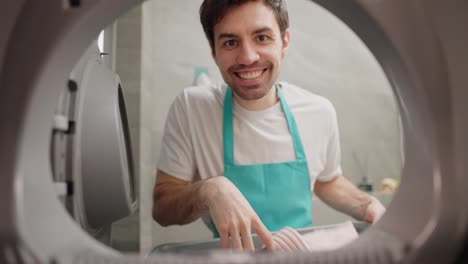 Portrait-of-a-creepy-brunette-janitor-a-man-in-a-white-T-shirt-and-blue-apron-slowly-closes-the-washing-machine-door-and-looks-inside-with-open-eyes-and-a-creepy-smile-while-cleaning-in-a-modern-apartment