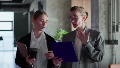 Two-businesswoman-girls-in-business-clothes-and-jackets-walk-along-the-corridor-in-a-modern-office-and-communicate-about-specific-paper-on-a-tablet