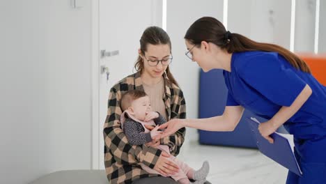 Over-the-shoulder-of-a-confident-brunette-girl-a-pediatrician-in-a-blue-uniform-a-doctor-playing-with-the-little-daughter-of-a-brunette-girl-in-a-checkered-shirt-in-a-modern-clinic