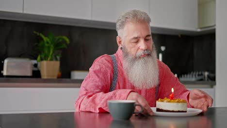 A-sad-elderly-man-with-gray-hair-and-a-lush-beard-in-a-pink-shirt-blows-out-a-candle-on-a-cake-during-his-lonely-birthday-party-in-a-modern-apartment