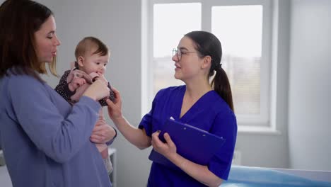 A-happy-brunette-girl-in-a-blue-uniform-communicates-with-a-doctor-along-with-her-little-child.-Childrens-pediatrician-and-doctor-for-children-communication.-Confident-brunette-girl-with-her-little-daughter-at-the-girl-childrens-doctor-in-a-modern-clinic