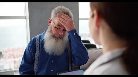 Por-Encima-Del-Hombro,-Un-Hombre-De-Pelo-Gris-Y-Gafas-Con-Una-Barba-Exuberante-Y-Una-Camisa-Azul-Se-Queja-Con-Una-Médica-Sobre-Sus-Problemas-Y-Dolores-De-Cabeza-Durante-Un-Examen-Domiciliario-En-Un-Apartamento-Moderno.