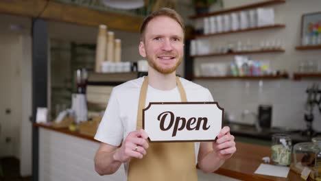 Porträt-Eines-Blonden-Kerls-Mit-Bart,-Der-In-Einem-Café-Ein-Schild-Mit-Einem-Offenen-Schild-In-Den-Händen-Hält-Und-Posiert