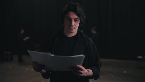 Portrait-of-a-confident-Happy-man-with-medium-length-brunette-hair-in-a-black-suit-an-actor-who-holds-a-piece-of-paper-in-each-of-his-hands-and-reads-the-text-of-your-character-and-the-script-of-the-play-before-preparing-to-perform-on-stage-in-the-theater