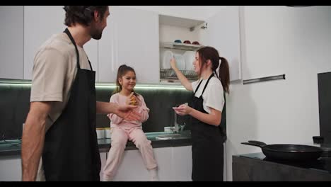 Happy-brunette-girl-in-a-white-T-shirt-and-black-apron-together-with-her-husband-with-brunette-stubble-and-little-daughter-in-pink-clothes-are-preparing-for-breakfast-and-laying-out-plates-and-food-in-the-morning-in-a-modern-apartment