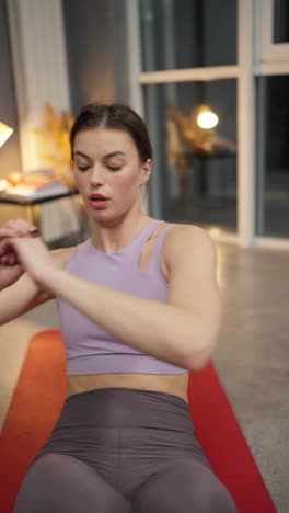 Vertical-video-close-up-of-a-confident-brunette-girl-in-a-purple-sports-summer-uniform-on-a-red-sports-mat-doing-sports-and-pumping-up-her-abs-in-a-modern-apartment-in-the-evening