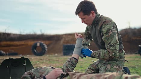 Side-view-of-a-confident-male-military-man-in-a-green-camouflage-uniform-with-medical-gloves-bandaging-the-hand-with-fabric-bandages-of-a-wounded-soldier-during-a-training-exercise-at-a-military-training-ground-with-a-barricade-of-wheel-tires