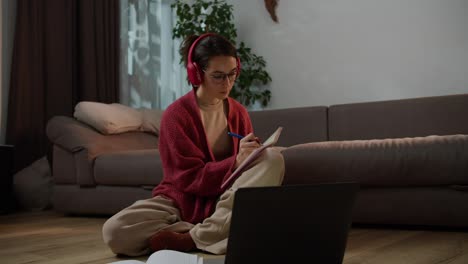 A-confident-and-serious-brunette-girl-with-glasses-in-wireless-red-headphones-and-a-sweater-sits-on-the-floor-in-front-of-a-gray-laptop-and-writes-down-her-notebooks-for-a-foreign-language-lesson-online