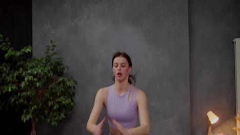 Confident-athletic-brunette-girl-in-purple-top-doing-squats-and-jumping-up-in-modern-apartment-while-doing-sports-at-home.-Confident-brunette-girl-doing-sports-at-home-in-a-modern-apartment-with-gray-walls