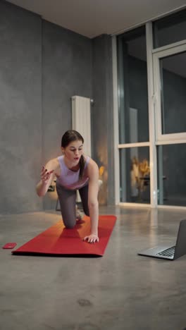 Vertical-video-of-a-confident-brunette-girl-in-a-sports-summer-uniform-doing-sports-on-a-Red-sports-mat-using-video-lessons-via-a-laptop-in-a-modern-apartment