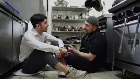 A-cheerful-guy-a-brunette-waiter-in-a-white-shirt-communicates-with-his-colleague-a-cook-in-a-black-uniform-during-a-break-at-work-in-a-restaurant-in-the-kitchen