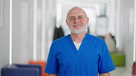 Portrait-of-a-happy-elderly-male-doctor-in-glasses-with-a-gray-beard-in-a-blue-uniform-standing-in-the-corridor-of-a-modern-light-clinic