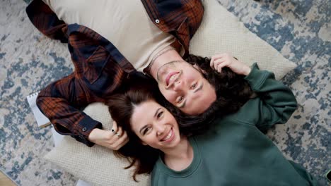 Top-view-rotating-camera-happy-brunette-girl-holding-and-touching-hair-of-her-boyfriend-with-curly-hair-lying-on-pillow-head-to-head-on-floor-in-modern-apartment-at-home