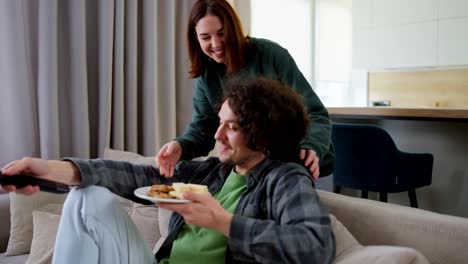 Happy-brunette-guy-with-curly-hair-communicates-with-his-girlfriend-who-brought-him-snacks-while-watching-a-movie-on-TV-at-home