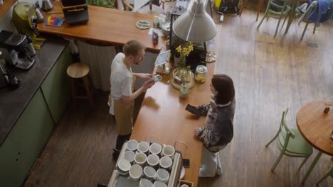 Top-view-of-a-blond-guy-in-an-apron,-a-cafe-worker-accepts-payment-from-a-brunette-girl-at-the-counter-in-a-cafe