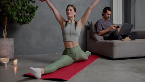 A-confident-brunette-girl-in-a-gray-top-made-of-green-pants-sits-on-the-splits-on-a-red-sports-mat-and-does-yoga-stretching-and-sports-near-her-her-boyfriend-who-is-doing-work-using-his-laptop-at-home-in-a-modern-apartment-near-the-burnt-candles-of-a-large-indoor-plant