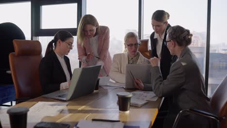 Confident-middle-aged-blonde-girl-with-glasses-in-a-white-business-suit-communicates-with-her-colleagues-and-listens-to-their-ideas-and-suggestions-about-work-in-the-office-during-a-meeting-with-a-wooden-table-in-a-modern-office