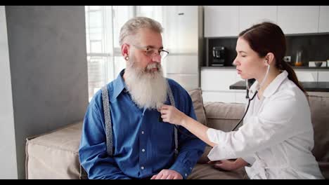 Un-Hombre-De-Pelo-Gris-Con-Gafas,-Barba-Exuberante-Y-Camisa-Azul,-Escucha-El-Veredicto-De-La-Novia-Del-Médico-Después-De-Un-Examen-Con-Un-Estetoscopio-En-Un-Apartamento-Moderno-En-Casa.