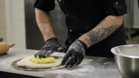 Close-up-a-cook-spreads-a-mixture-of-cheese-onto-a-prepared-flatbread-of-dough-for-making-a-cheese-flatbread-on-a-table-on-which-there-is-flour-and-kitchen-utensils-for-baking-in-a-restaurant