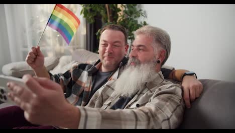 Happy-couple-of-middle-aged-LGBT-men-a-brunette-in-a-plaid-shirt-and-a-man-with-gray-hair-and-a-lush-white-beard-are-sitting-on-a-sofa-and-taking-a-selfie-in-a-modern-apartment-with-an-LGBT-flag