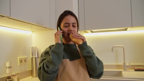 A-happy-and-confident-brunette-girl-in-a-green-jacket-and-a-beige-apron-eats-a-croissant-while-communicating-with-her-loved-ones-using-a-green-phone-and-rejoices-in-a-modern-apartment-in-the-kitchen