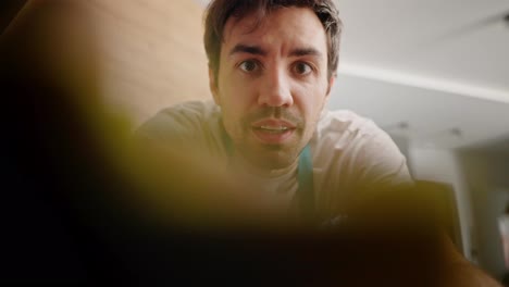 Confident-brunette-guy-with-stubble-in-a-white-T-shirt-and-blue-apron-wipes-the-camera-with-a-yellow-rag-and-poses-with-it-while-cleaning-the-kitchen-in-a-modern-apartment