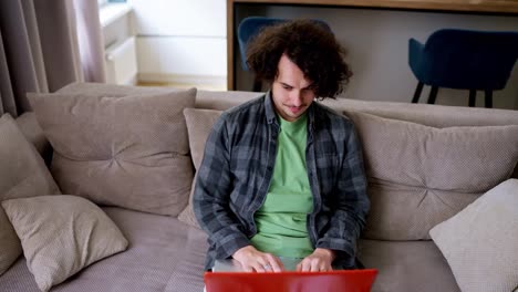 Happy-brunette-guy-with-curly-hair-in-a-gray-checkered-shirt-sits-on-a-light-brown-shirt-at-a-laptop-in-a-modern-apartment